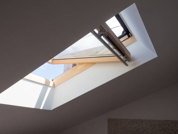 Dormer wooden window in the white sloping ceiling overlooking the blue sky. Sunlight enters the room through a opened window